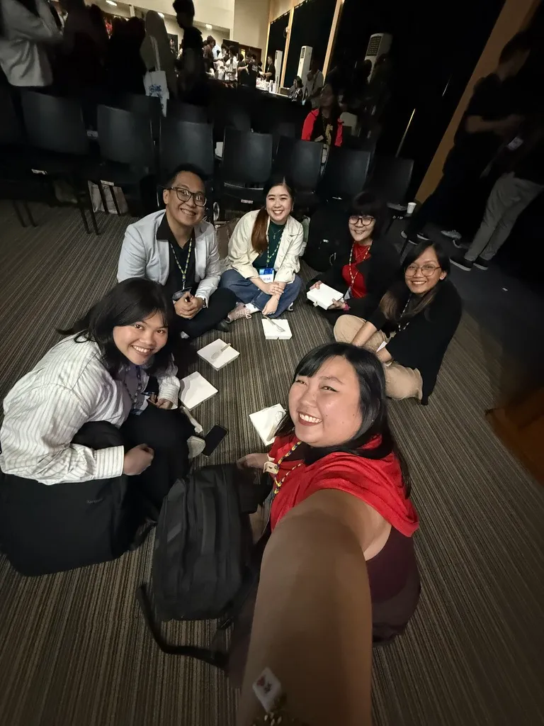 Chi doing a wide angle selfie with other workshop facilitators and speakers from UXPH Mini 2025: Davao. They are seen sitting on the floor of an auditorium while eating lunch in a group huddle setup.