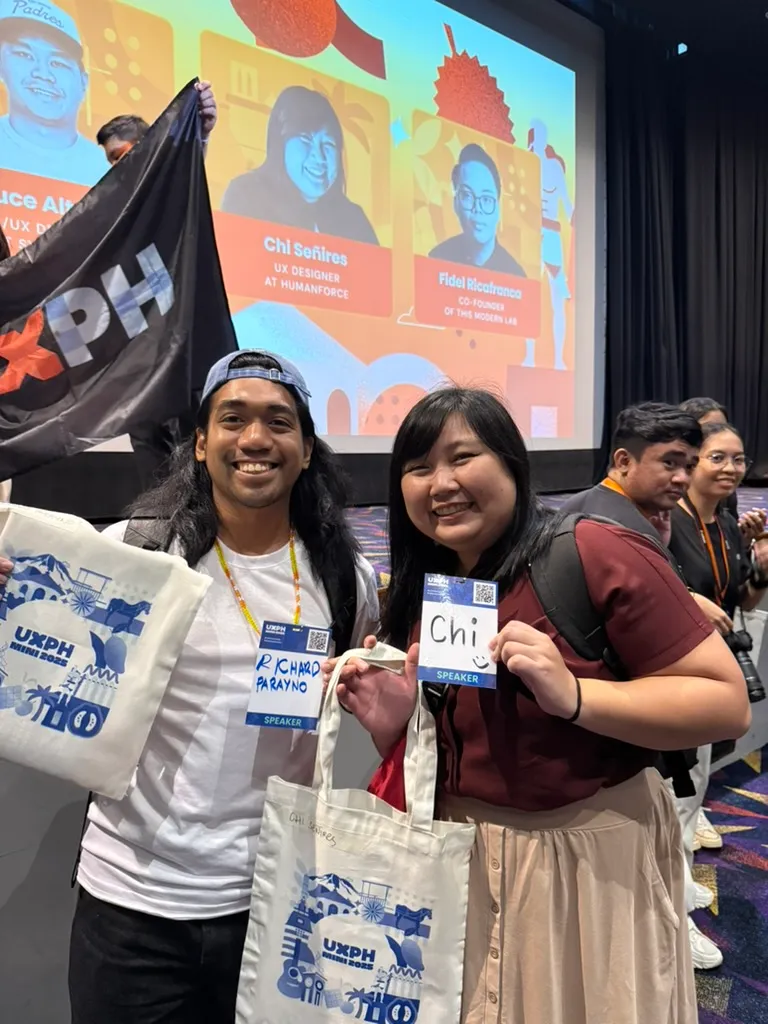 Richard and Chi posing near the stage for UXPH Mini 2025: Cebu, with both of them holding up their conference ID tags and their swag bags with the UXPH Mini branding printed on it.