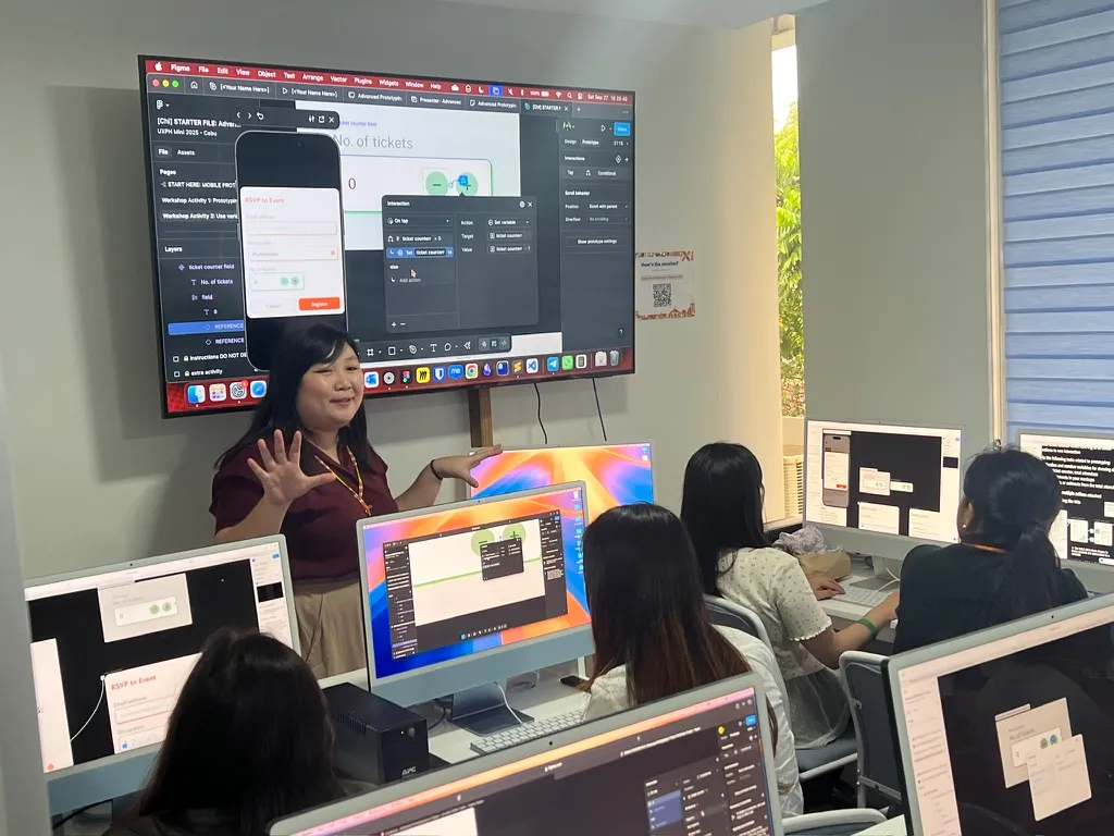 Chi is standing in front of a small classroom, both her hands up as she explains what is shown on the big TV screen behind her, which is sharing her laptop’s screen with Figma open and the prototype preview overlay showing a modal within a phone frame. Attendees are positioned around and in front of her, with iMacs on each table and attendees also following along in Figma during the activity.