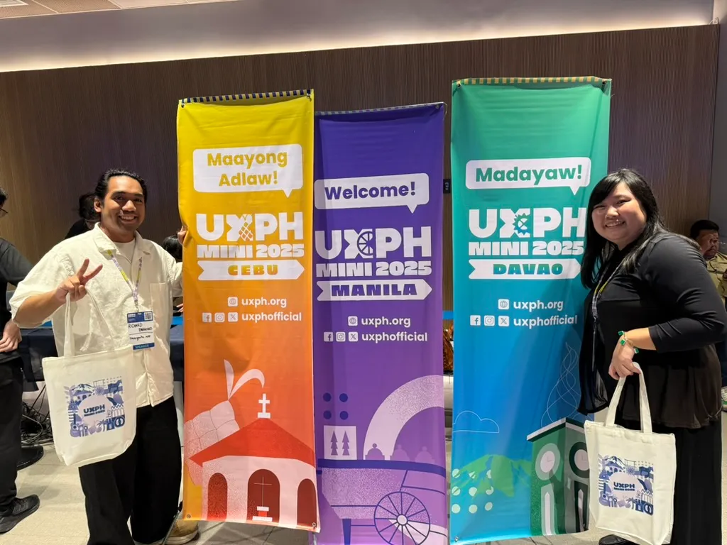 Chi and Richard posing in front of the 3 tarps for the different legs for UXPH Mini 2025. Richard is by the left side of the Manila leg tarp holding up a peace sign, and Chi is on the right side with the Davao leg tarp. Both Chi and Richard are also holding on to their conference swag bags.