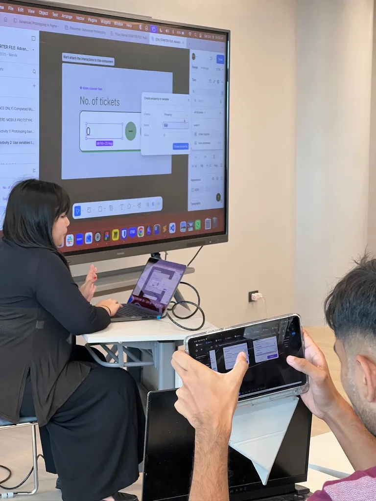 Chi is seated in front of a classroom, gesturing to her laptop that is projecting her screen, showing her fiddling with some prototyping interaction settings on Figma. To the right of the photo, an attendee is holding up their iPad that has Figma open, following along the workshop activities.