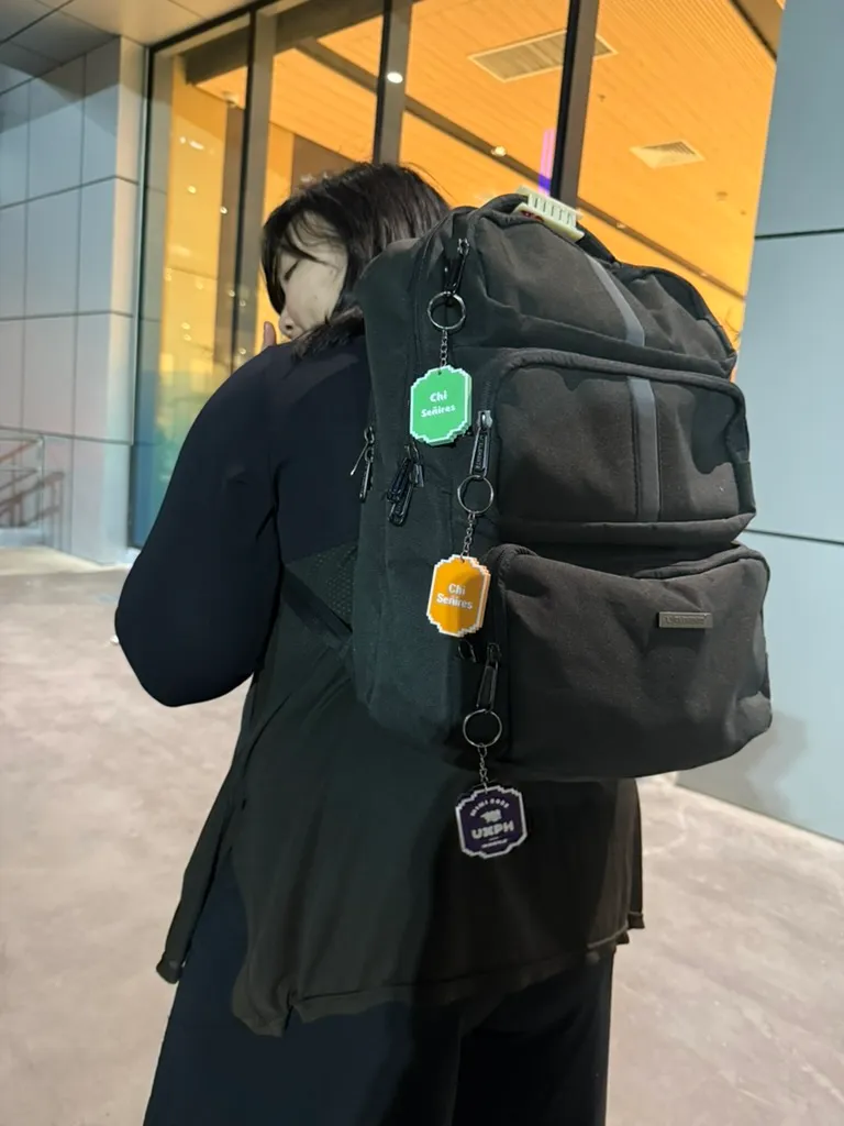 Chi holding up her backpack that has 3 distinct 3d printed keychains on 3 different zippers, showing off the different conference tokens from the UXPH Mini 2025 legs. The top one is a green keychain for the Davao leg, the middle one is an orange keychain for the Cebu leg, and the bottom one is a purple keychain for the Manila leg.