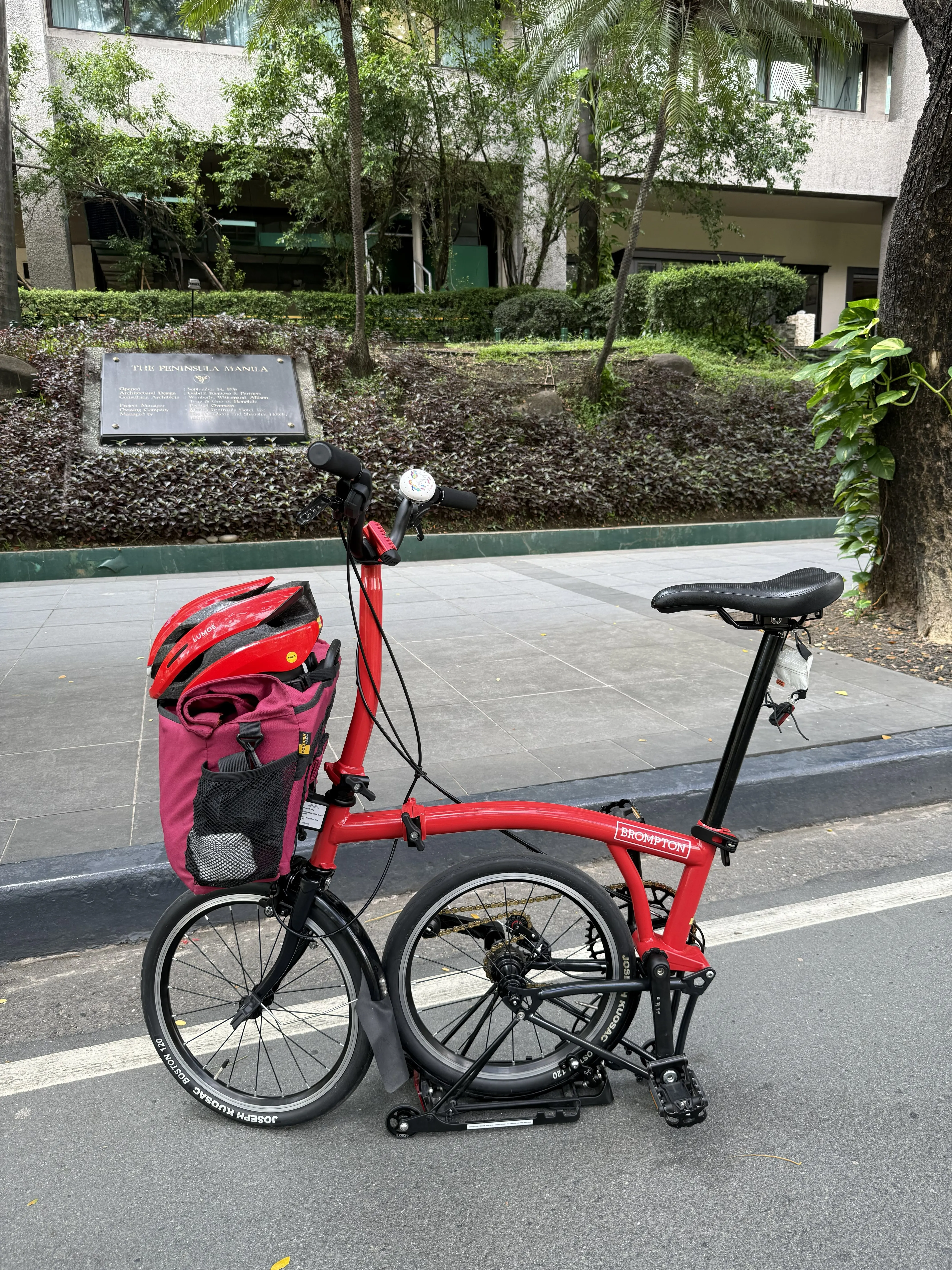 A red Brompton bike with its rear wheel folded under, allowing it to stand upright on its own. A red bag is mounted on the front rack with a matching red helmet resting on top. The bike is parked on the roadside in front of The Peninsula Manila, whose plaque is visible behind some greenery.